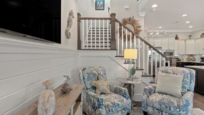 A living room with two patterned armchairs, a wooden bench, a starfish decoration, a television, and a staircase leading to a kitchen. - Three Times The Fun by AvantStay | Blue Mountain | Pool + Pet-Friendly (Santa Rosa Beach)