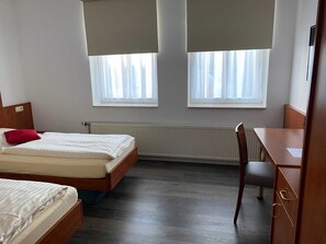Quarto com duas camas, uma escrivaninha e uma cadeira. Possui duas janelas com persianas, um radiador e uma porta.