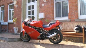 A red motorcycle parked in front of a brick building with a number 45 sign, a bench, and a potted plant.