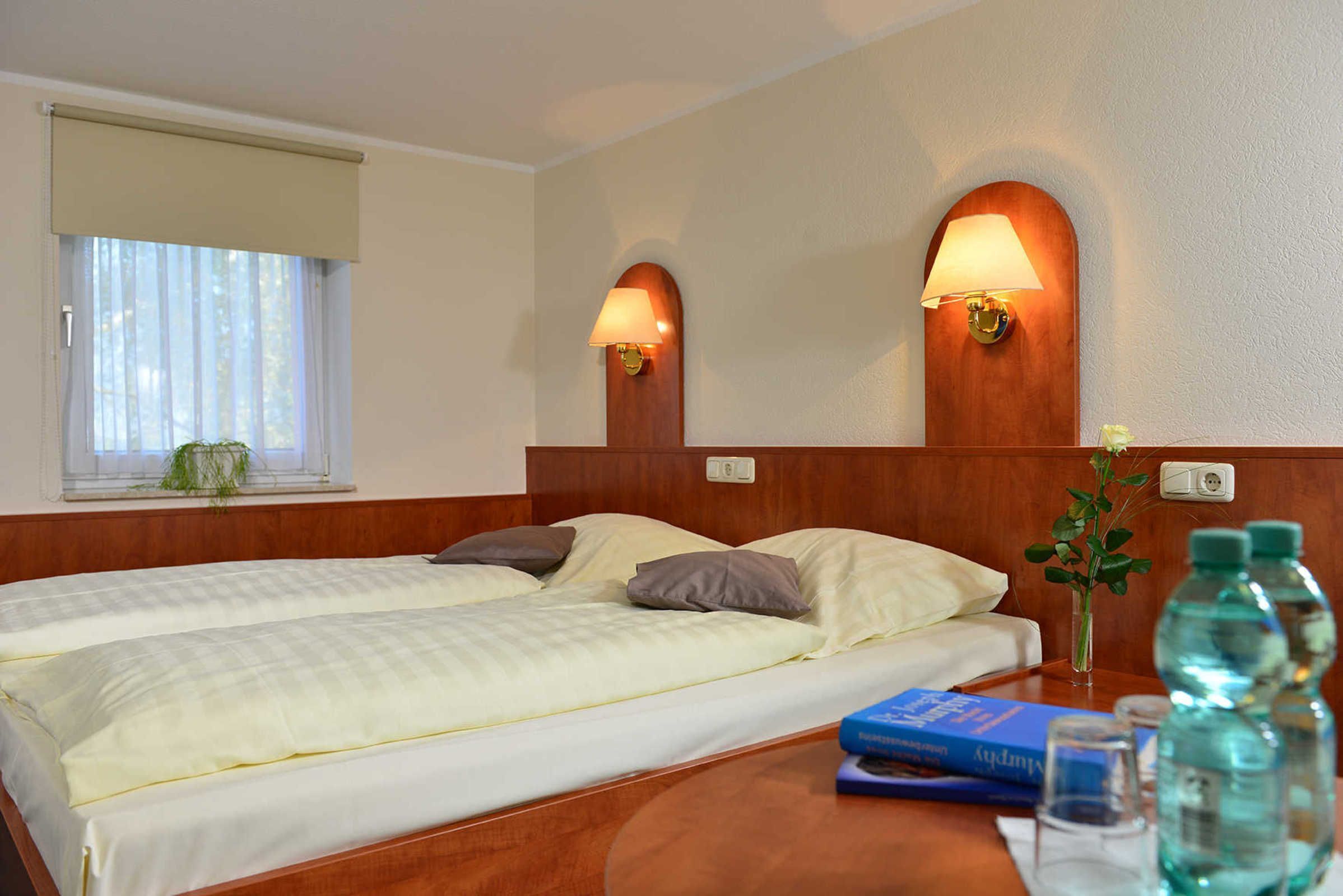 A hotel room with two beds, a wooden headboard, two wall-mounted lamps, a window with a blind, a small table with a vase of flowers, and a water bottle.