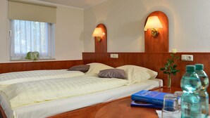 A hotel room with two beds, a wooden headboard, two wall-mounted lamps, a window with a blind, a small table with a vase of flowers, and a water bottle.