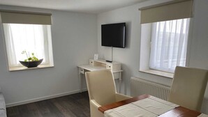 A modern room with a wooden dining table, a white desk, a television mounted on the wall, and a window with a plant on the windowsill.