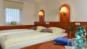 Hotellihuone, jossa on kaksi sänkyä, puinen pääty, ikkuna, jossa on sälekaihtimet, sekä yöpöytä, jossa on lamppu ja vesipulloja.
