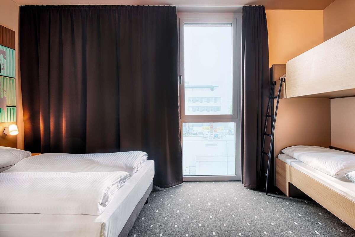 A hotel room with two beds, a window with curtains, a view of the street, and a small table with a lamp.