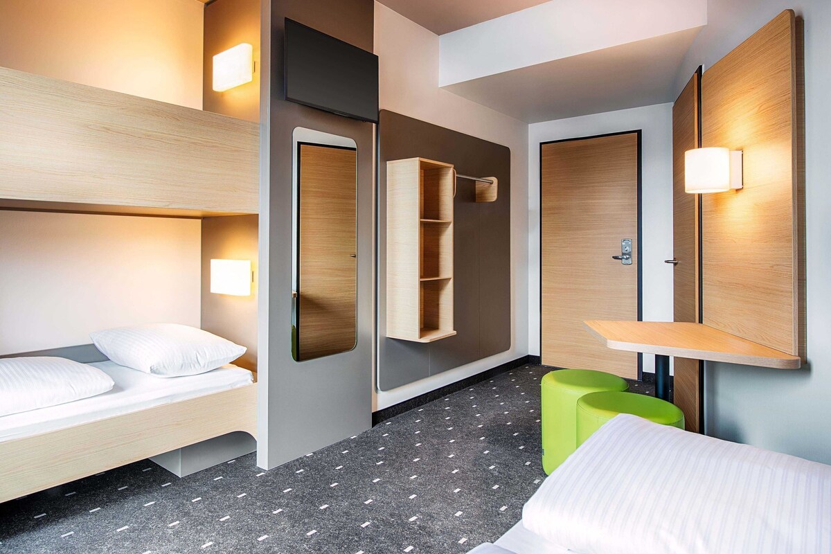 A modern hotel room with a bunk bed, a bedside table, a mirror, and a small green stool.