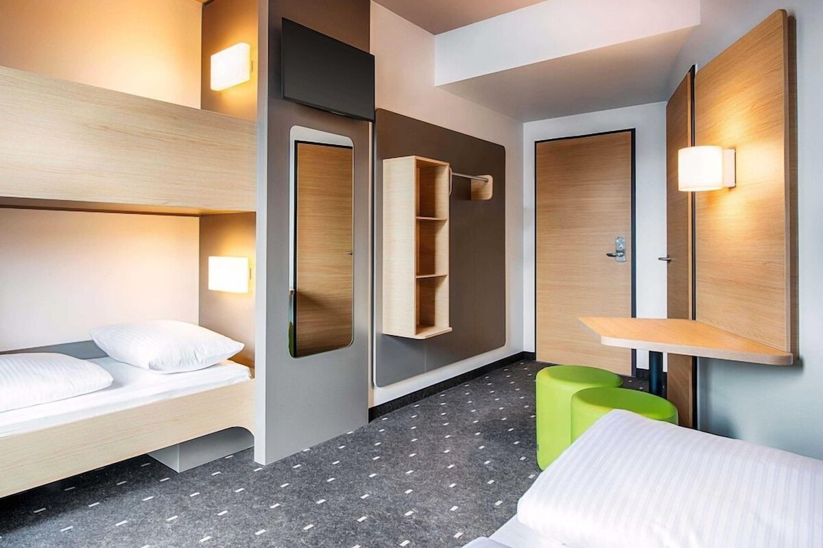 A hotel room with a bunk bed, a desk with a chair, a green stool, and a wall-mounted shelf.