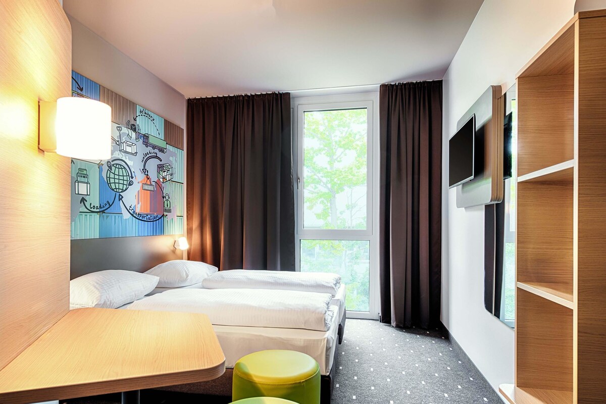 A hotel room with a bed, a desk, two green ottomans, a wall-mounted TV, and a window with curtains.