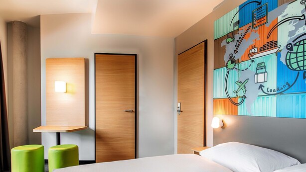 A hotel room with a bed, two green stools, a wooden wardrobe, and a wall with a colorful map and loading icon. - B&B Hotel Stuttgart-Neckarhafen (Stuttgart)