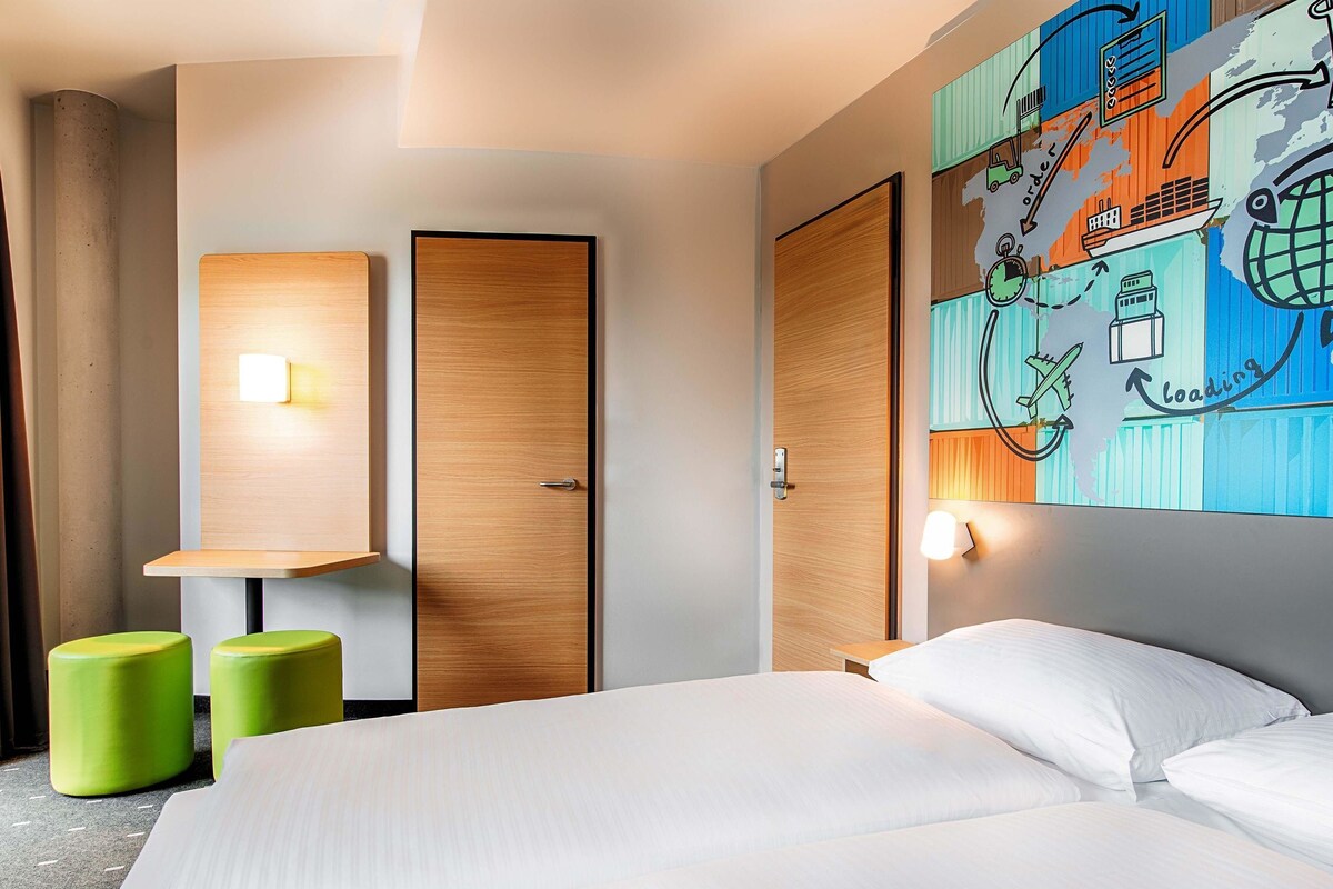 A hotel room with a bed, two green stools, a wooden wardrobe, and a wall with a colorful map and loading icon.