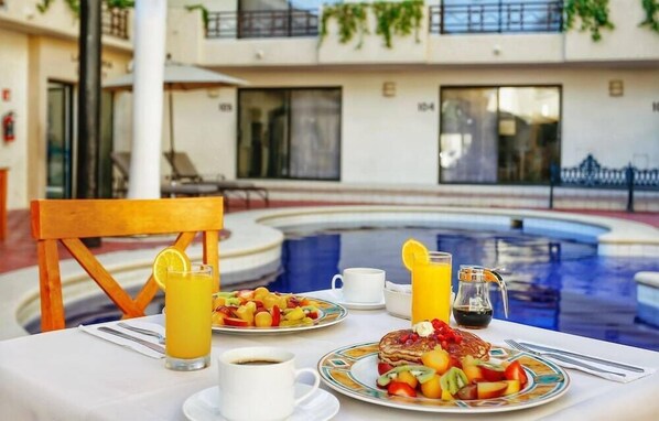 Una mesa de desayuno con platos de fruta, panqueques y café, con vista a la piscina y un edificio de hotel.