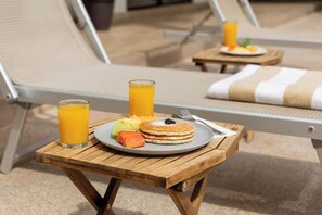 Una mesa de madera con alimentos para el desayuno, como panqueques, fruta y jugo, en una terraza soleada con sillones de descanso.