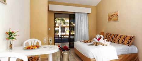 A hotel room with a bed, a dining table with chairs, a small table with a vase of flowers, and a ceiling fan.