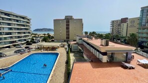 A hotel with a swimming pool, a covered area with sun loungers, and a view of the sea.