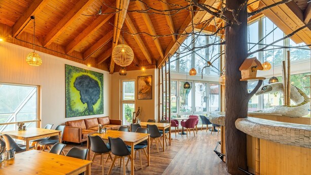 A restaurant with wooden tables and chairs, large windows, and a high ceiling with exposed beams.