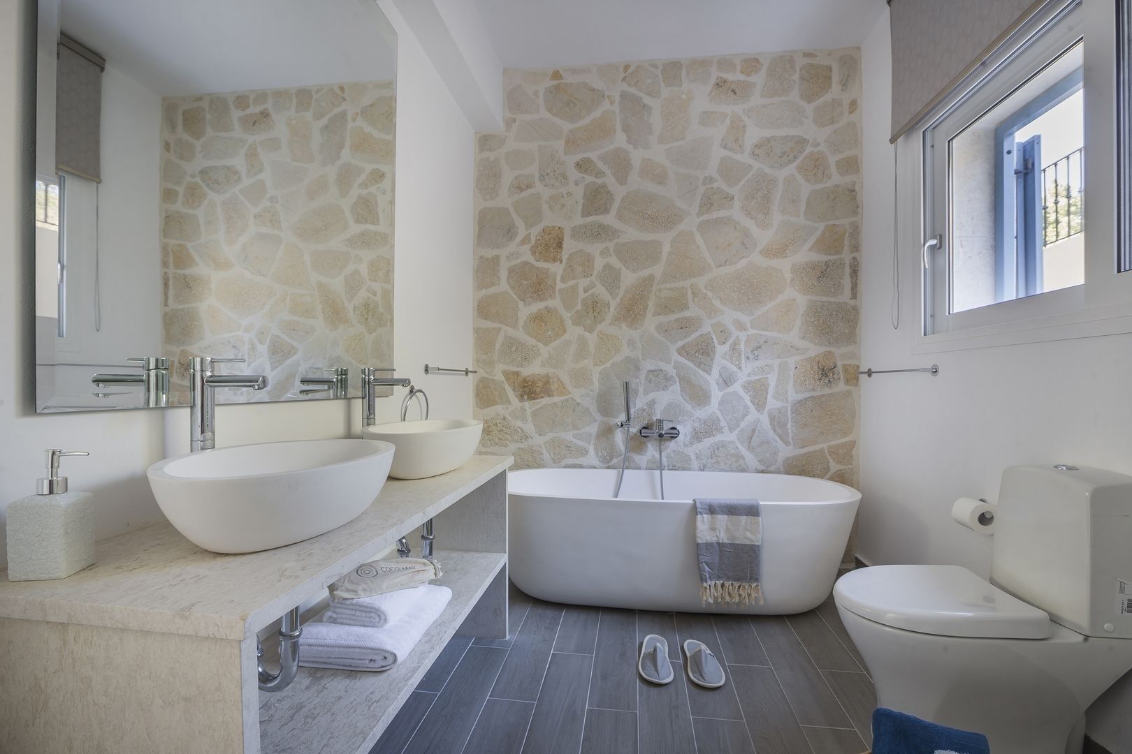 Un baño con pared de piedra, dos lavamanos, una bañera y un inodoro.