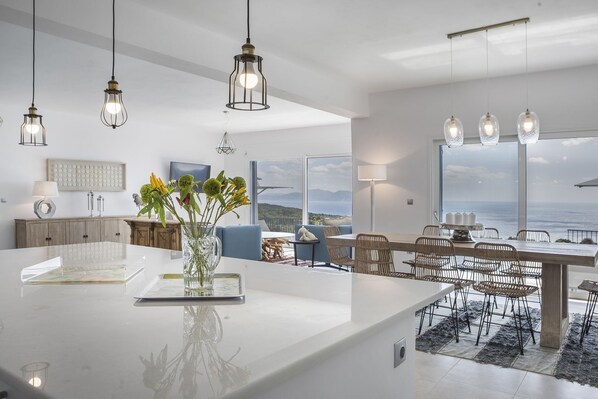 Une cuisine moderne dotée d’un grand îlot central, d’un coin repas avec des chaises en osier et offrant une vue sur l’océan.
