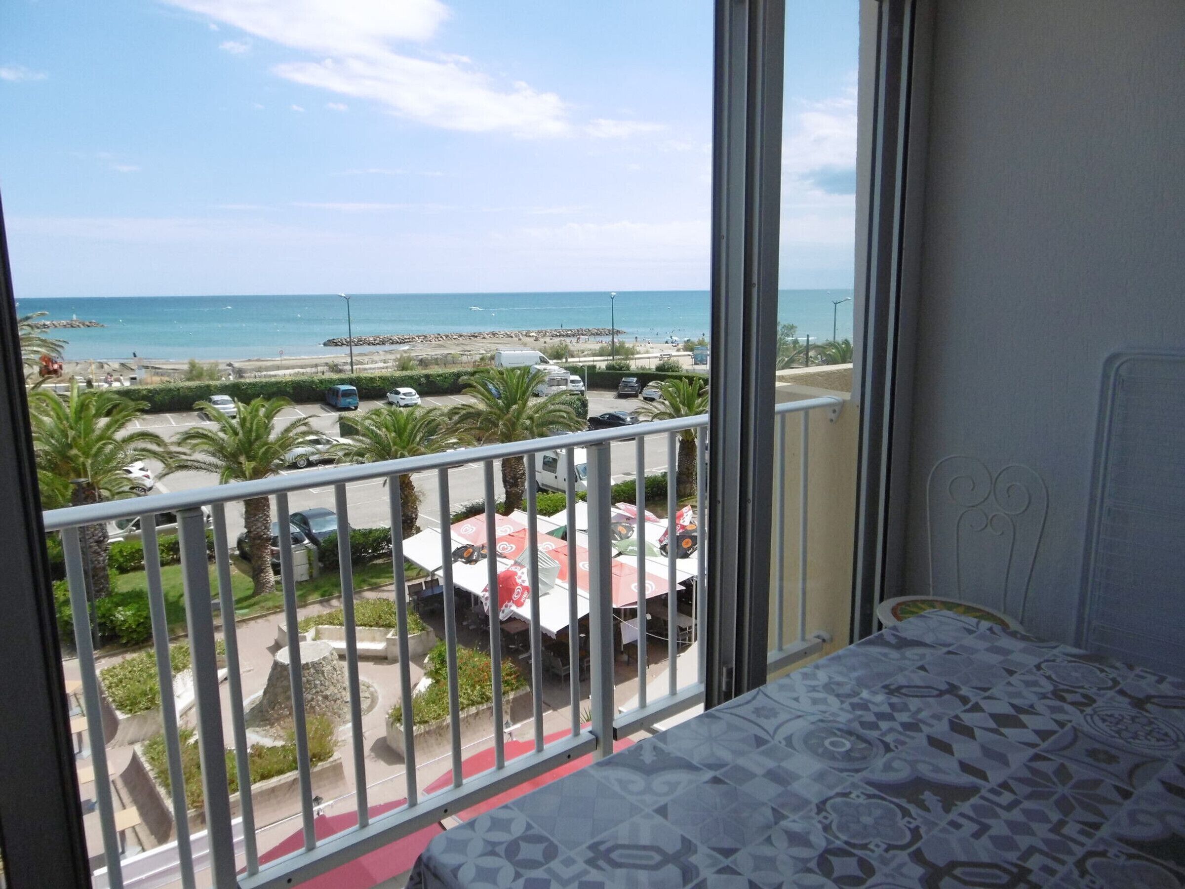 A balcony with a view of the beach, parked cars, and a dining area.