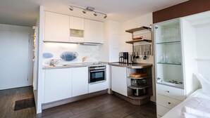A compact kitchen with white cabinets, a built-in oven, and a glass-fronted cabinet with kitchenware. - App. Kröger 42, Deutschland (Sylt)