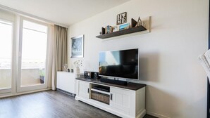 A living room with a flat-screen TV on a white entertainment unit, a large window with curtains, and a view of the outdoors. - App. Kröger 42, Deutschland (Sylt)