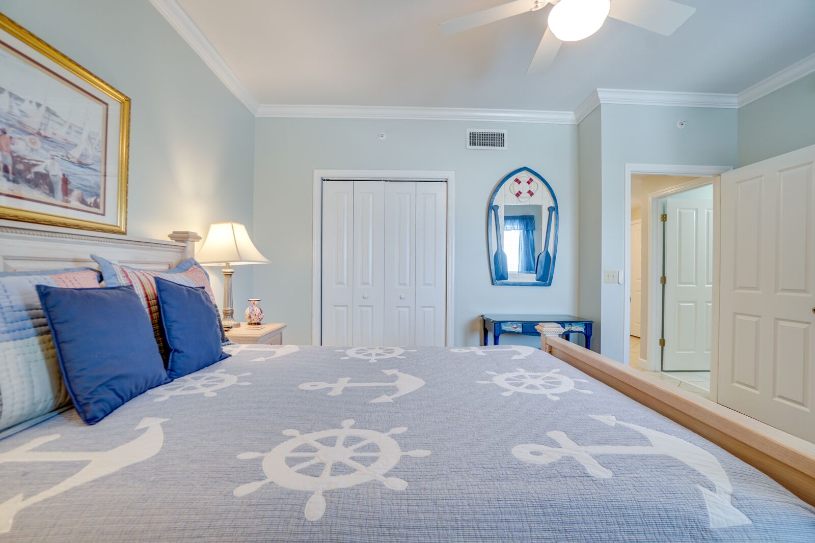 Een slaapkamer met een bed, voorzien van een beddegoed met een nautisch thema, een nachtkastje met een lamp, een dressoir en een spiegel aan de muur.