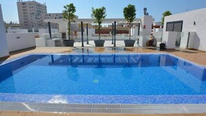 En pool på taket med kristallklart blått vatten, omgiven av vita väggar och planterade blommor i krukor.