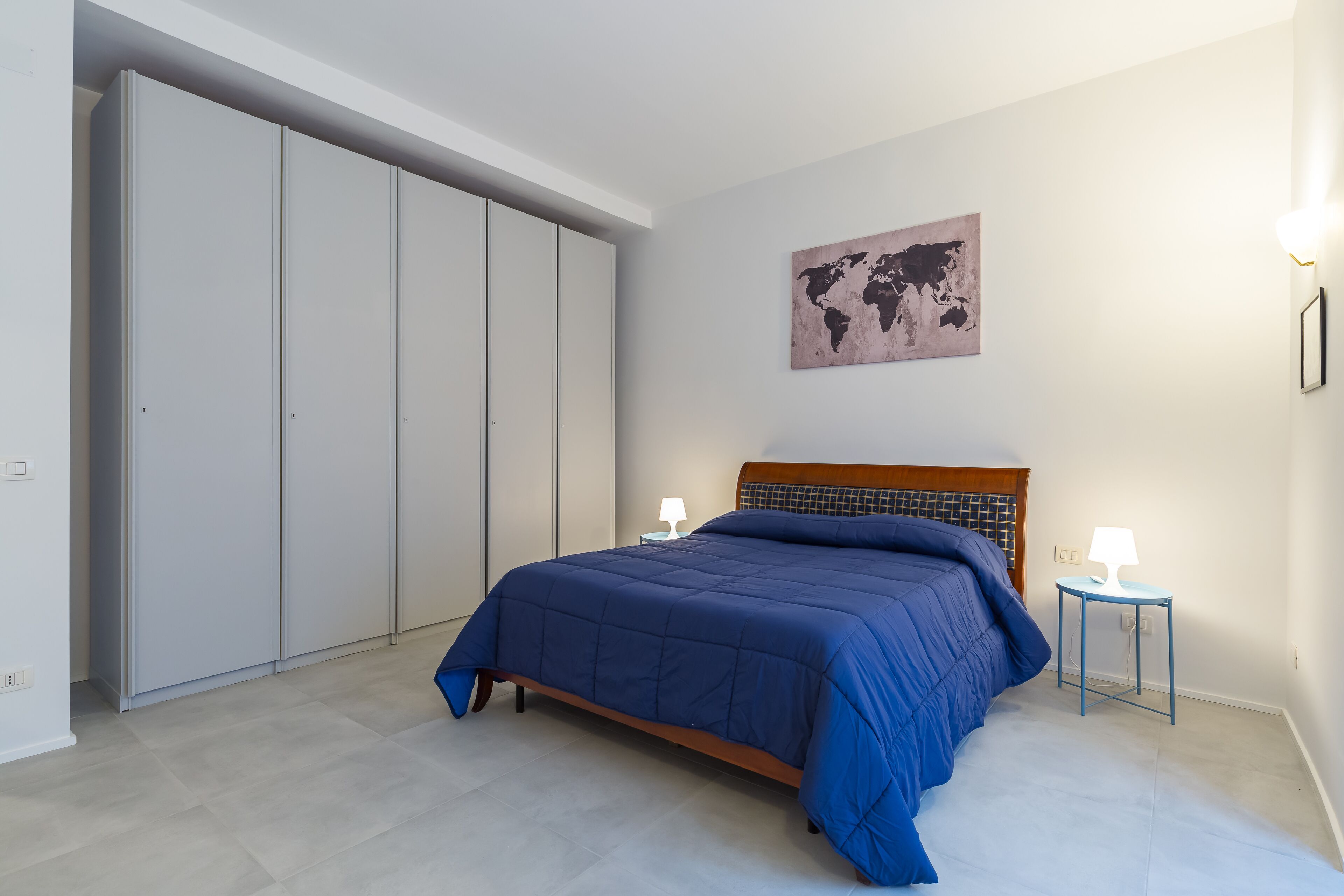 A bedroom with a bed, a wardrobe, a world map on the wall, and two bedside lamps.