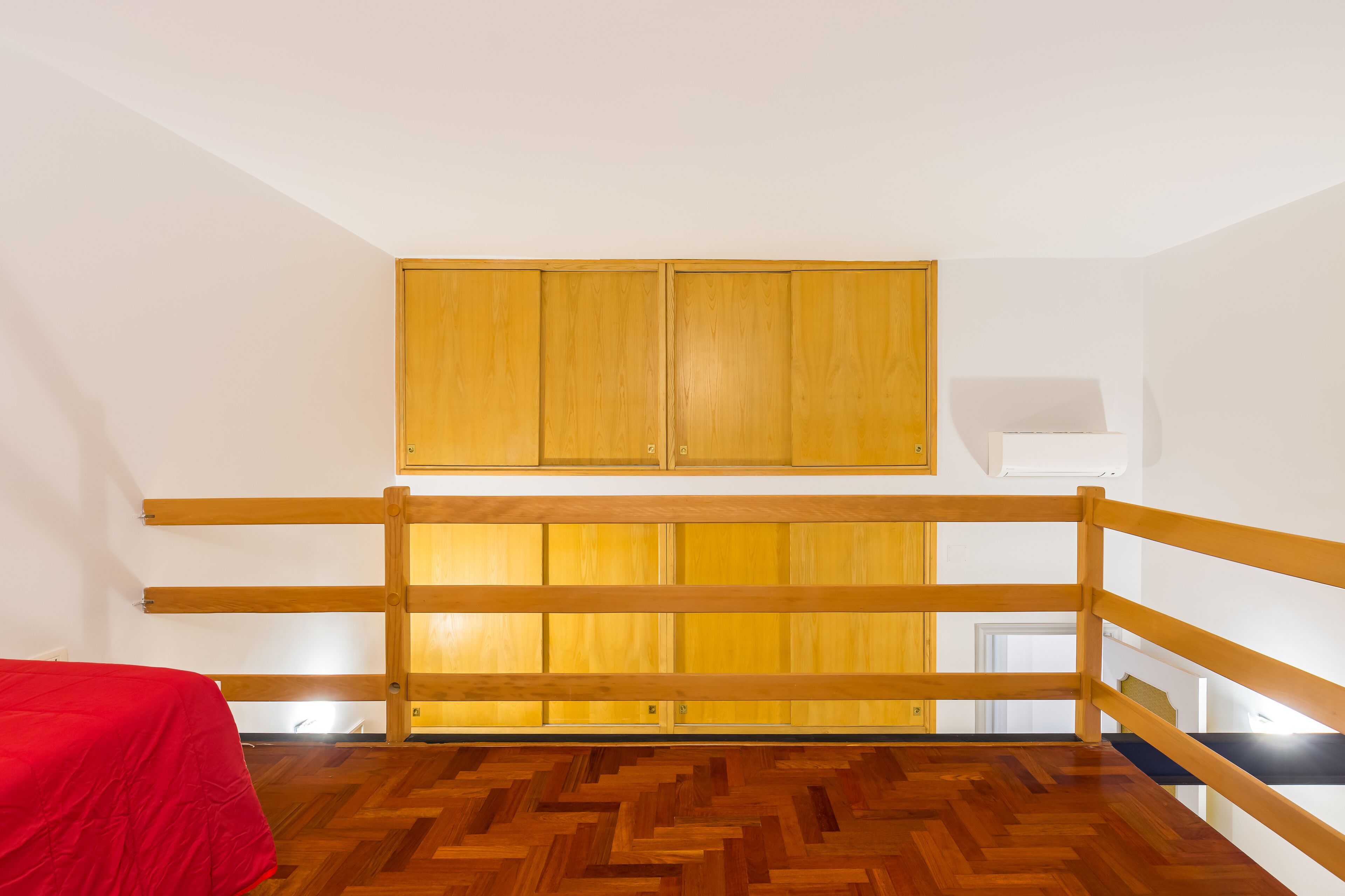Une chambre avec un lit superposé en bois, une couvre-lit rouge et un parquet en bois à motifs chevrons.