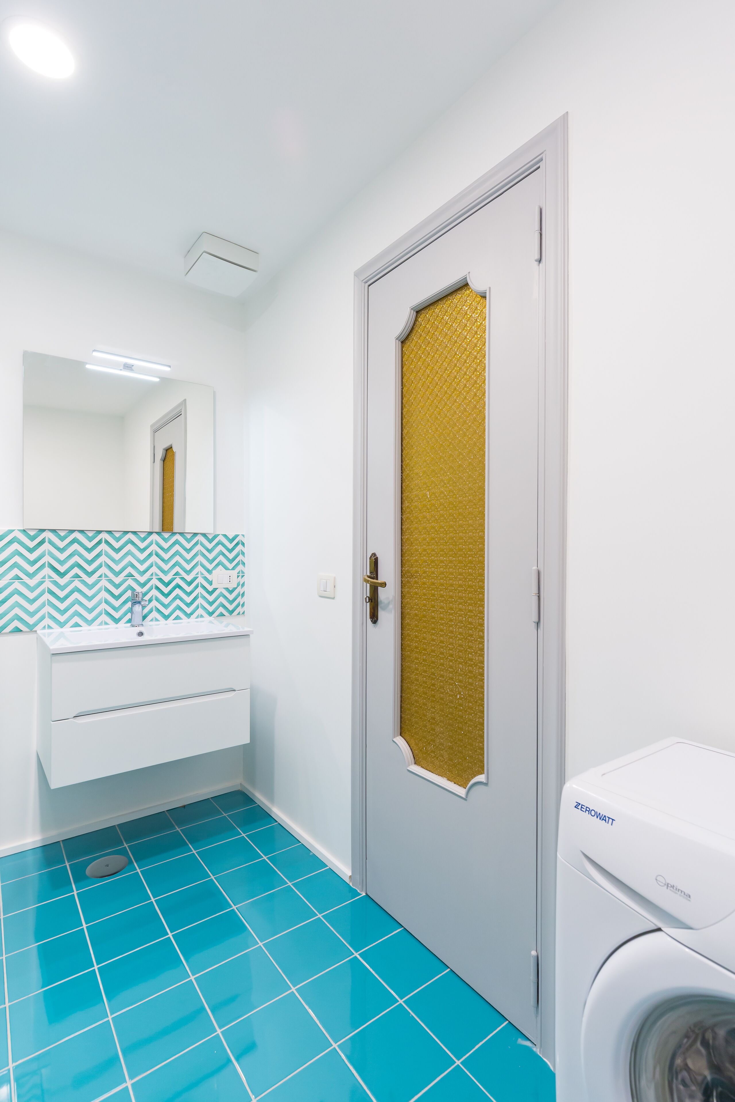 A bathroom with a white sink, a mirror, a blue tiled floor, a gold-patterned door, and a white washing machine.