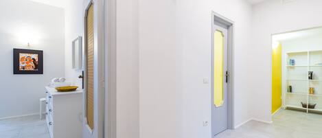 A hallway with white walls and a light-colored floor, leading to a room with a white cabinet and a framed picture on the wall.