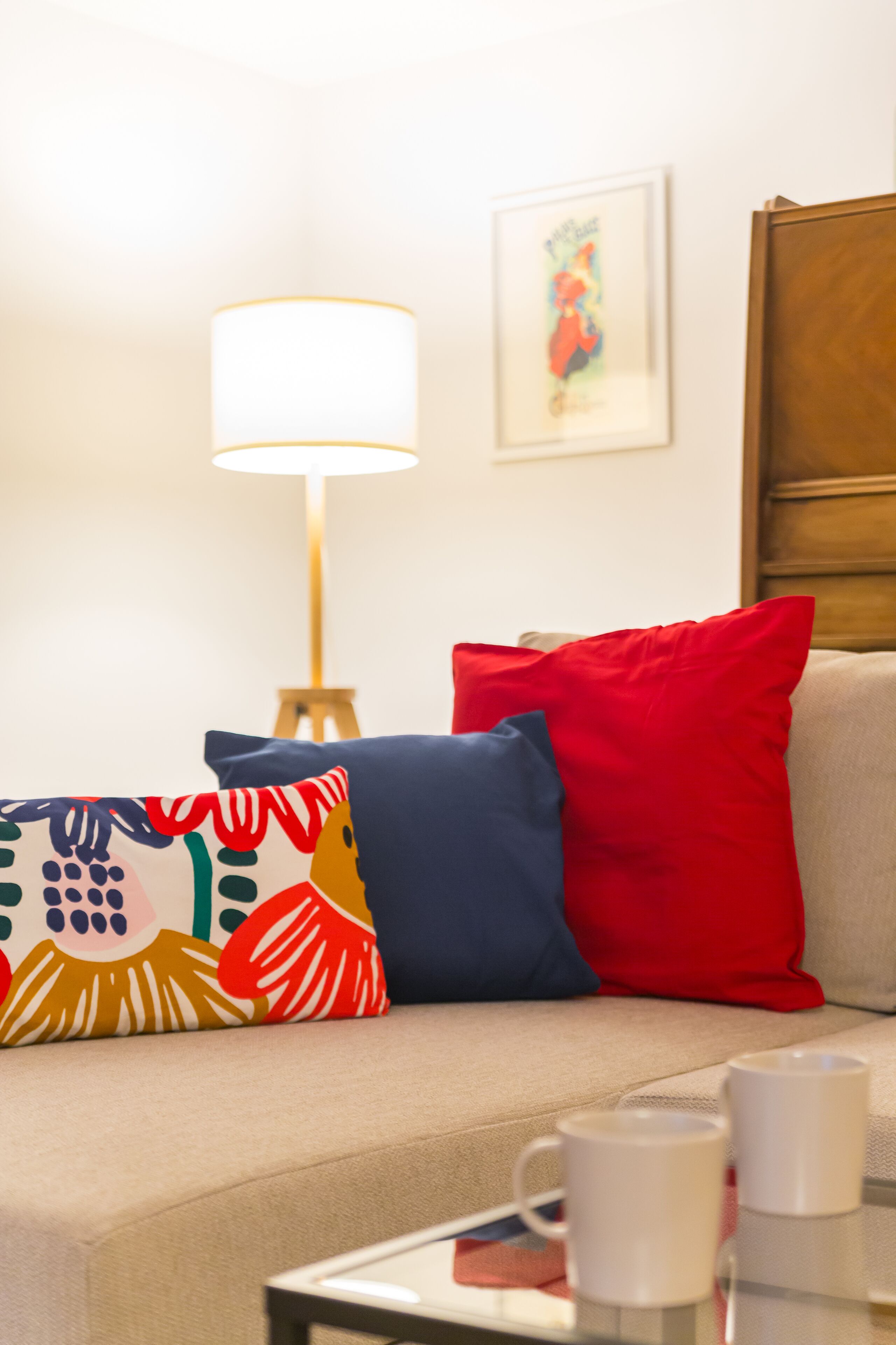 A living room with a beige sofa, colorful pillows, a white lamp, and a framed picture on the wall.