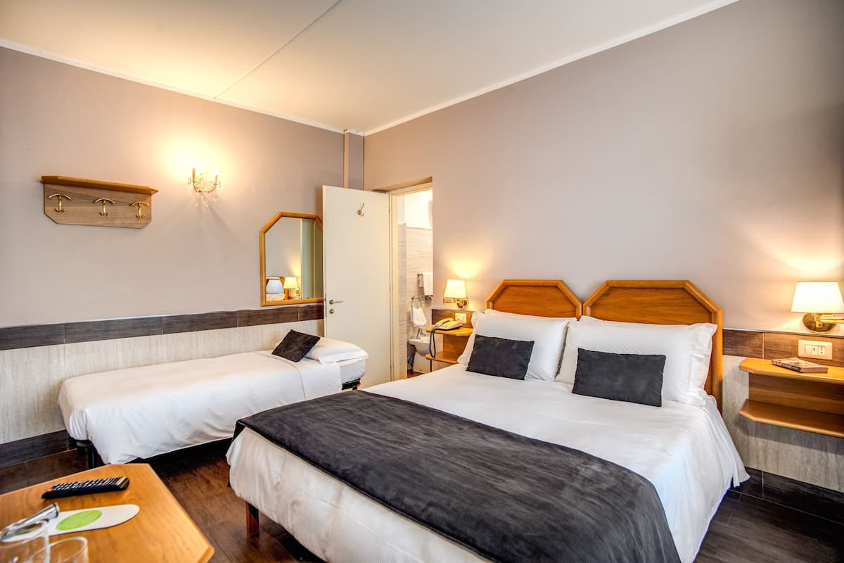 A hotel room with two single beds, a large bed, a wooden headboard, bedside tables with lamps, a mirror, a small table with a remote control, and a telephone.