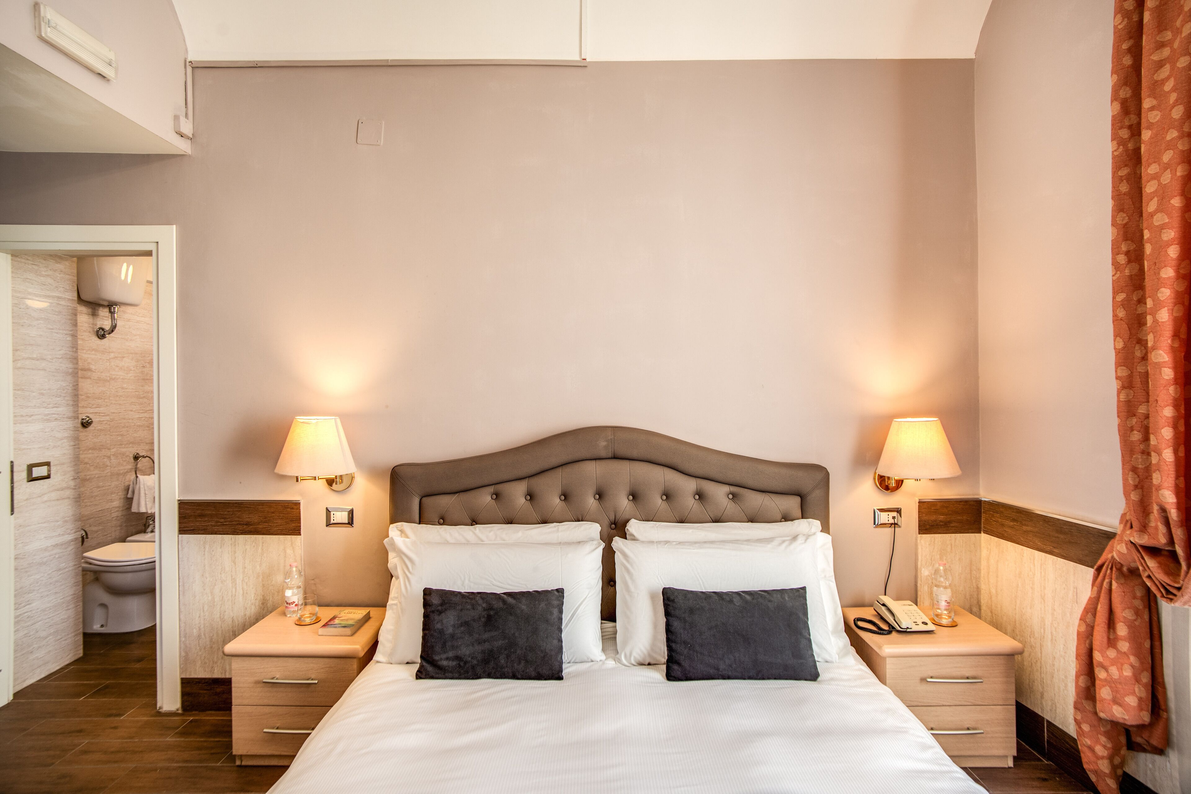 A neatly arranged hotel room with a large bed, two bedside tables, a nightstand, a lamp, and a bathroom visible through an open door.
