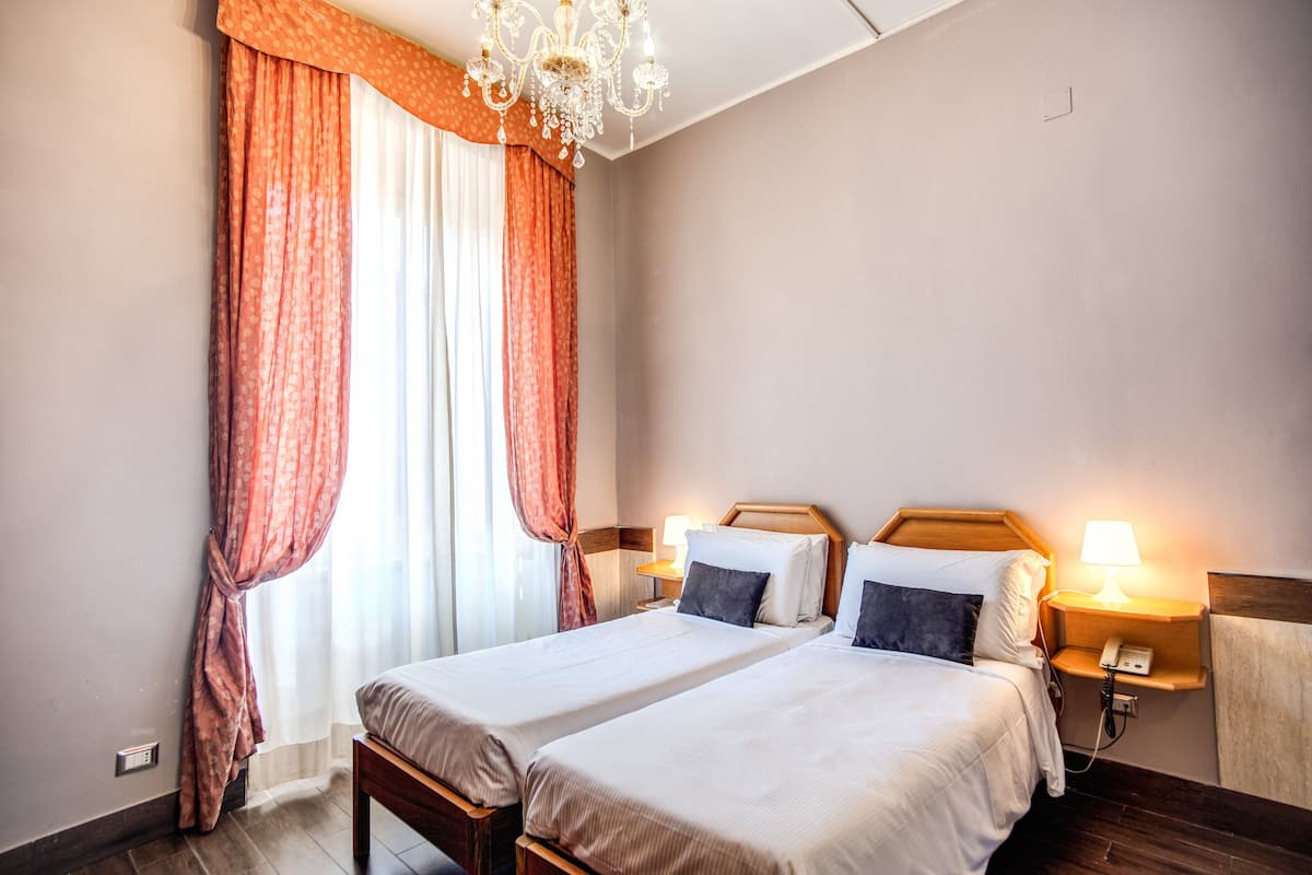 A hotel room with two beds, a chandelier, a nightstand with a lamp, and a window with curtains.