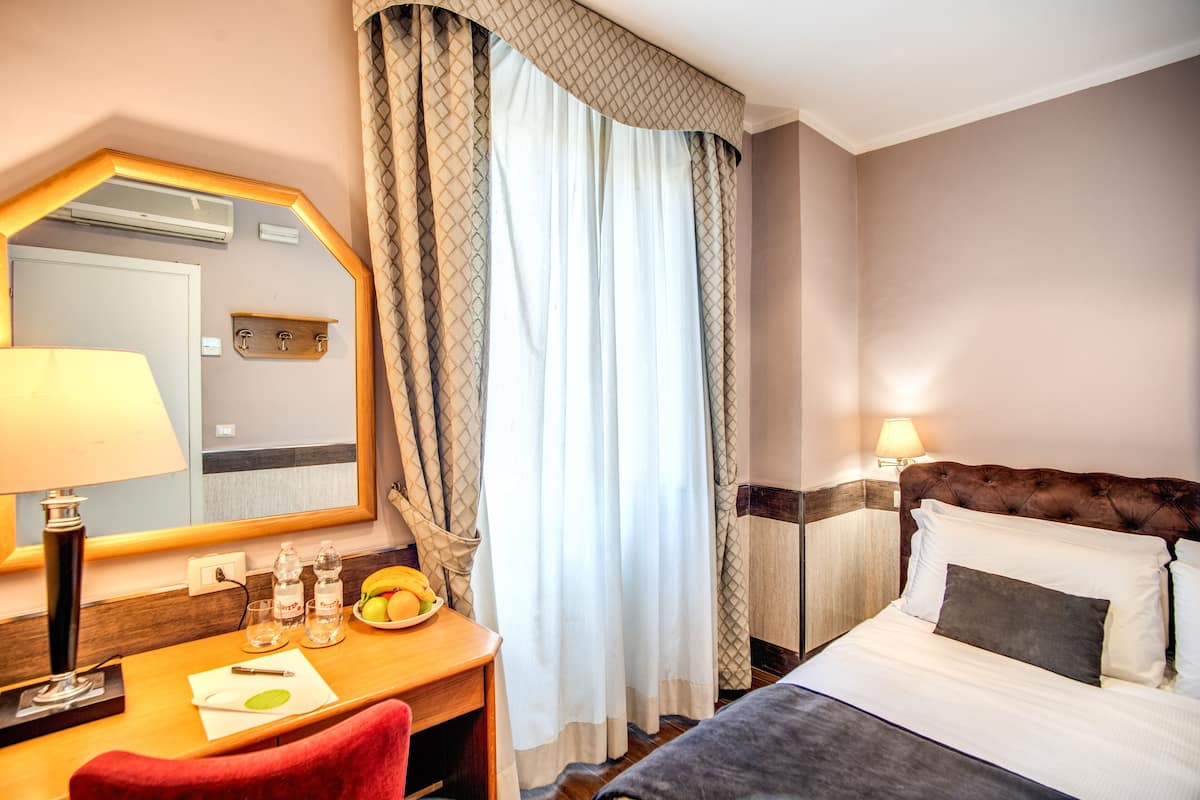 A hotel room with a bed, a desk with a lamp, a mirror, and a window with curtains.