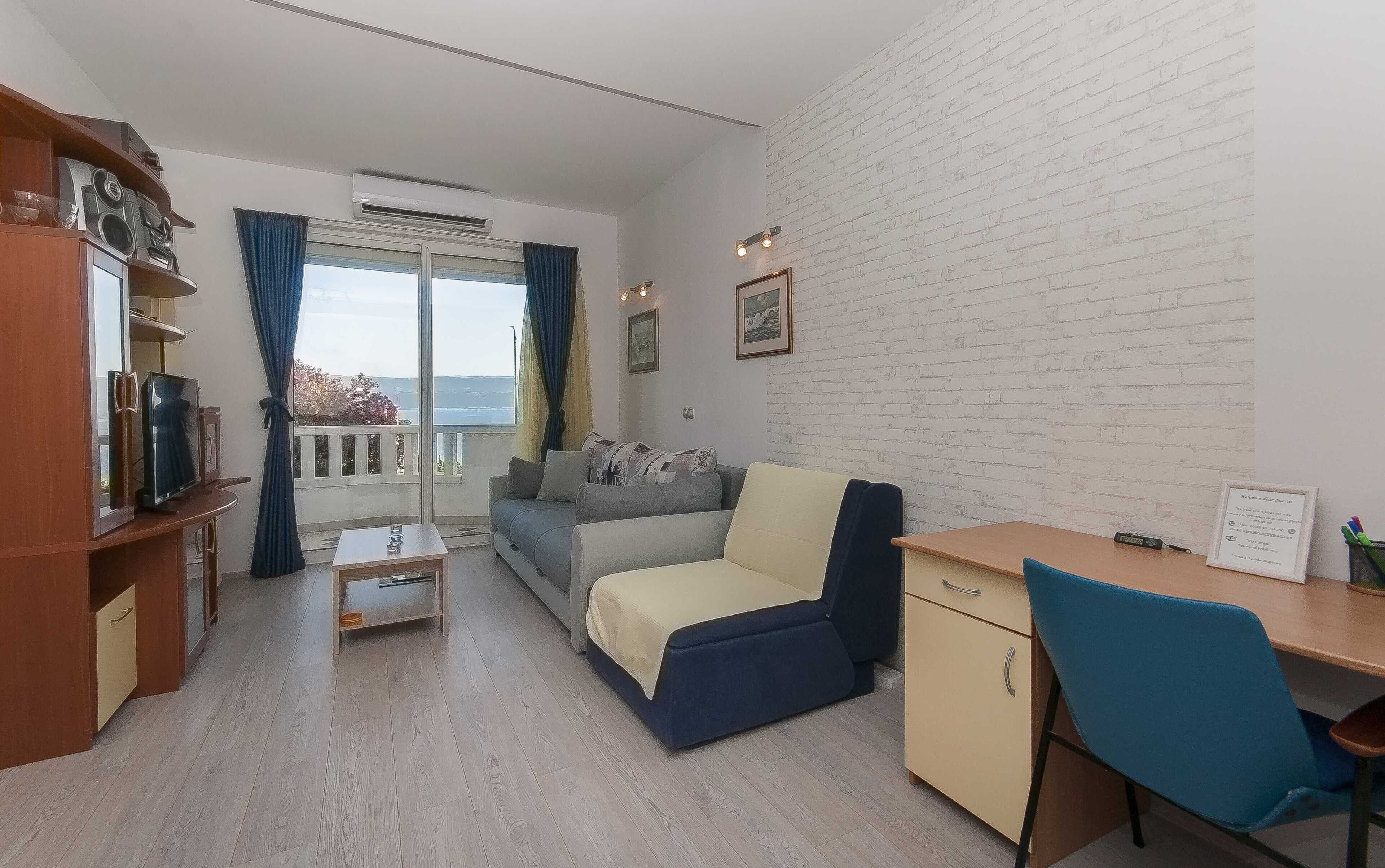 A compact living space with a sofa, a small table, a desk, and a cabinet. There is a window with curtains, a white brick wall, and a view of the outdoors.