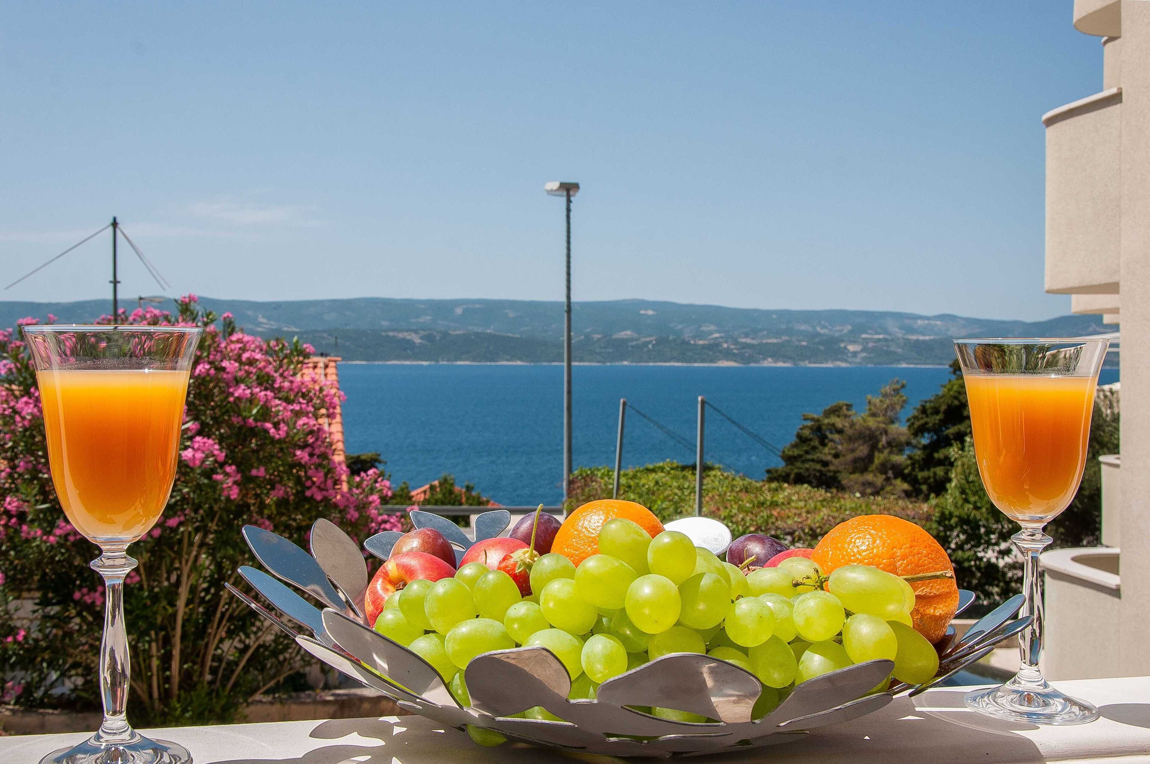 Una mesa con una cesta de fruta y dos vasos de zumo de naranja, con vistas a la costa.