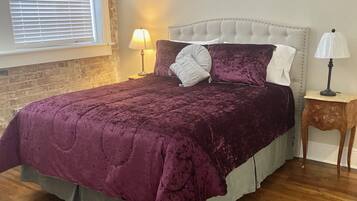 A bedroom with a large bed, two bedside tables, a window with blinds, and a brick wall.