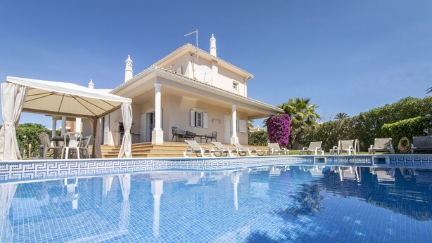 A swimming pool with lounge chairs and a covered area, a building with a tiled roof, and a clear blue sky.
