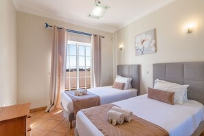 A hotel room with two beds, a window with curtains, a painting on the wall, and a wooden desk.