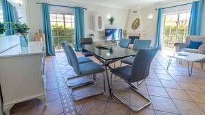 A modern living room with a glass dining table, four chairs, a sofa, and a TV.