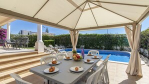 A covered outdoor dining area with a pool view, white chairs, and a table set with plates and cutlery.