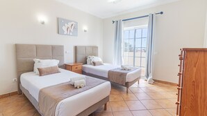 A hotel room with two beds, a nightstand, a wooden wardrobe, a window with curtains, and a wall-mounted light fixture.