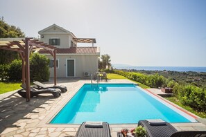 A swimming pool with lounge chairs and a pergola, a house with a tiled roof, and a view of the sea.
