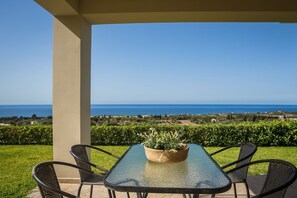 A glass-top table with four chairs, a potted plant, and a view of the sea and landscape.