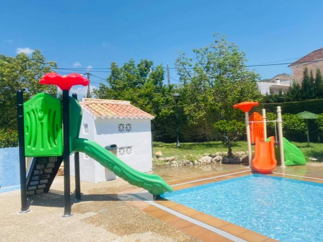 A swimming pool with two slides, one green and one orange, and a small white building with a tiled roof.