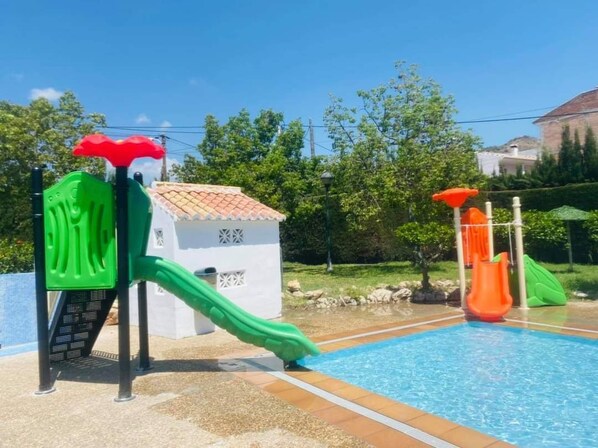 A swimming pool with two slides, one green and one orange, and a small white building with a tiled roof.