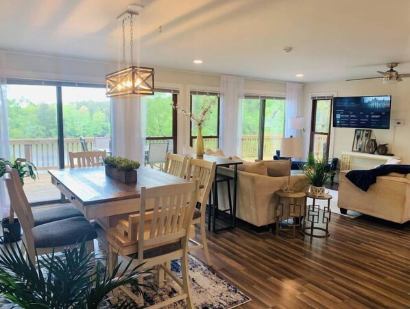 Relax in this inviting living area with a dining table, sofa, and TV, surrounded by large windows offering a view of the outdoors.