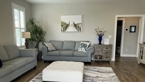 Un salon avec un canapé gris, un pouf assorti, un tapis à motifs, une table basse en bois, une table d’appoint avec une lampe, une plante en pot et un tableau accroché au mur.