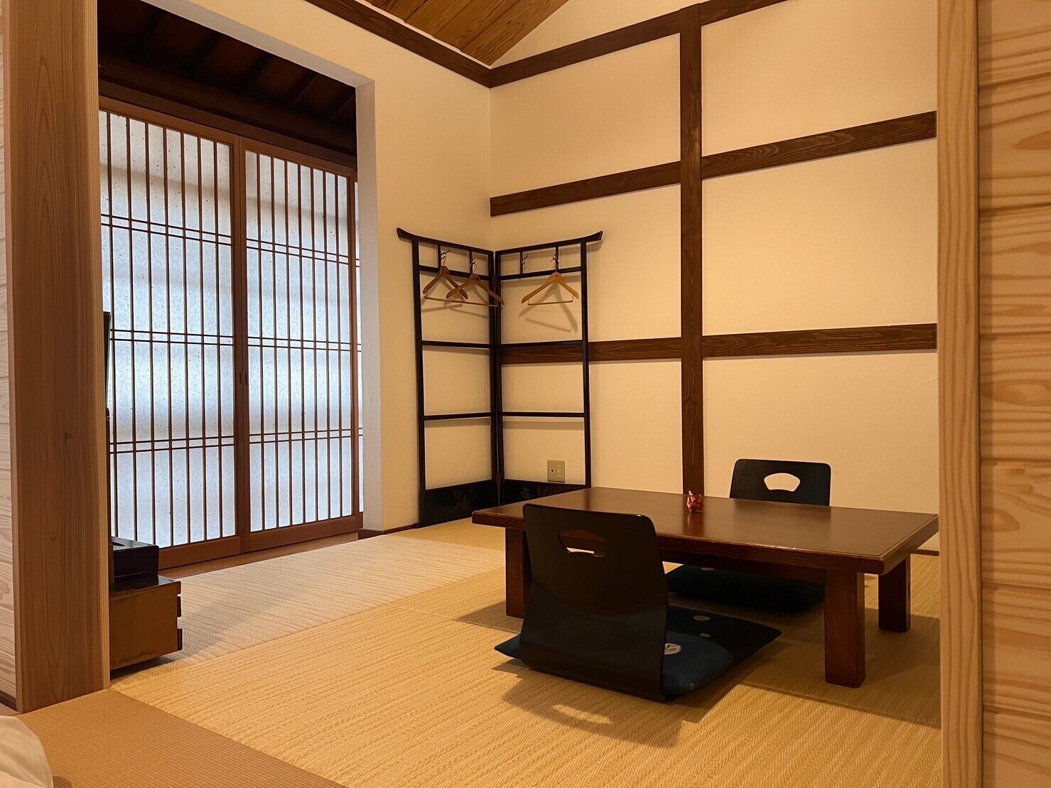 A traditional Japanese room with tatami flooring, a low table, a chair, a shoji screen, and a wooden shelf with hangers.