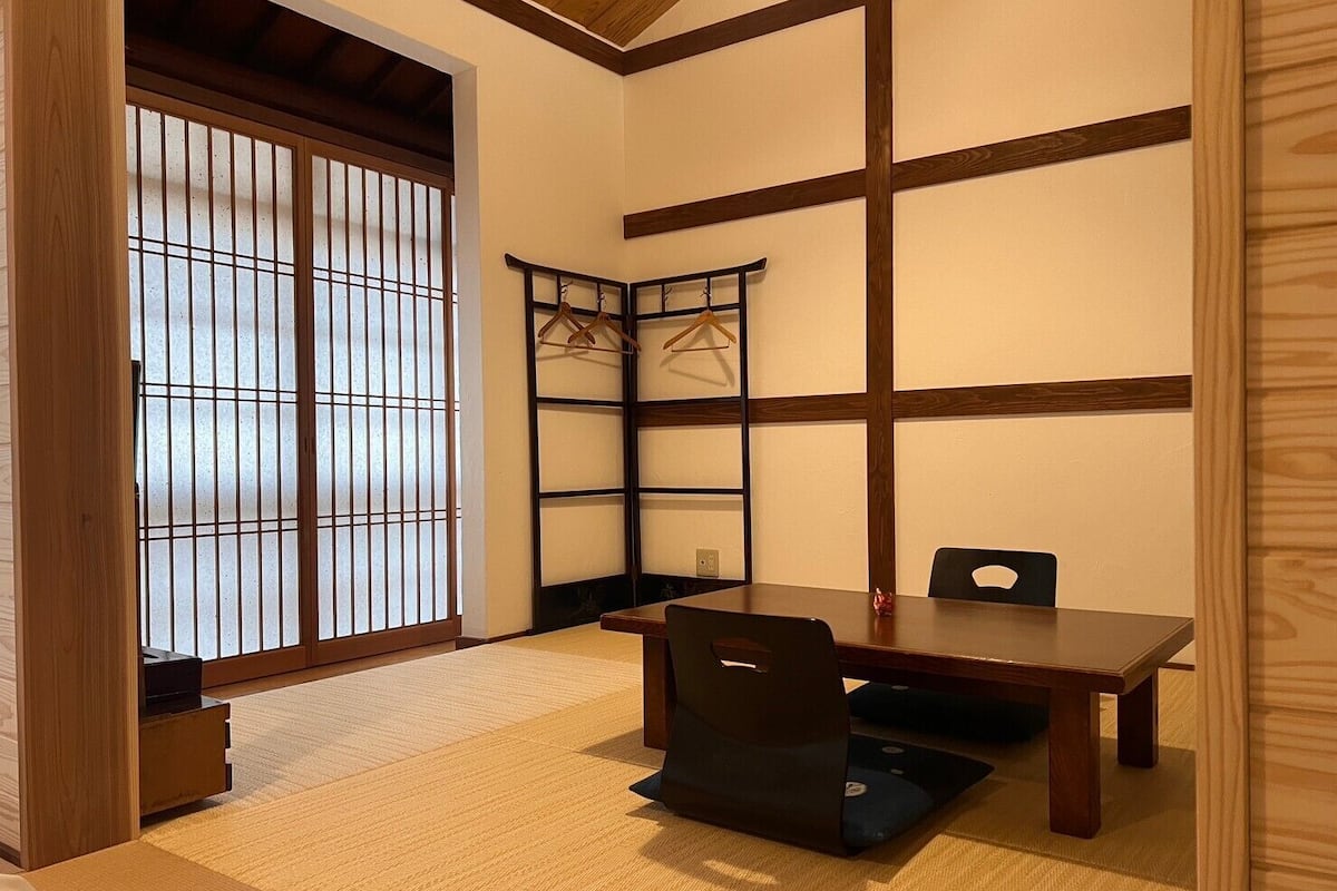 A traditional Japanese room with tatami flooring, a low table, a chair, a shoji screen, and a wooden shelf with hangers.